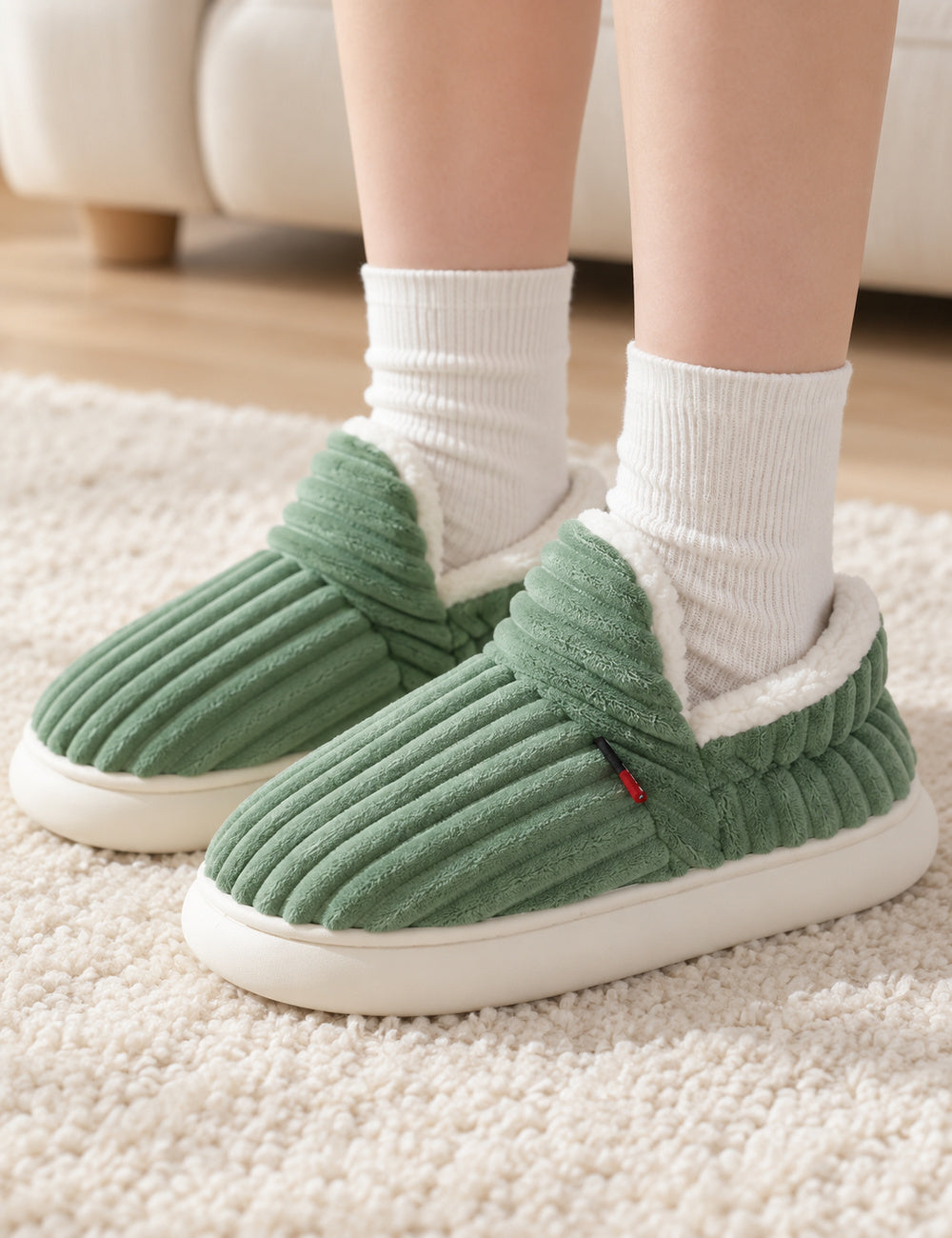 Chaussons Alix vert sauge portés avec des chaussettes blanches, pieds posés sur un tapis